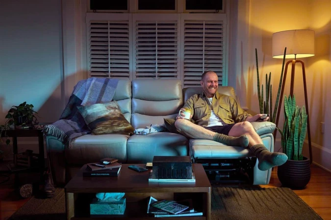 Man relaxing on a recliner sofa in a cozy living room at night, holding a mug and remote control, with soft lighting.