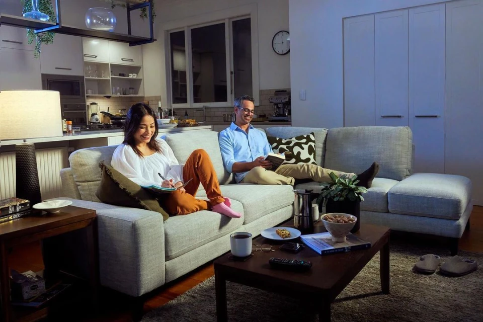 Couple relaxes on a gray sofa in an open-plan living room with modern decor, reading a book and writing in a notebook.