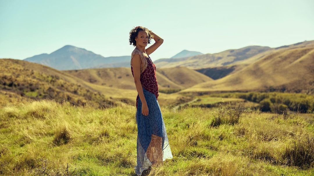 Woman with curly hair in a floral top and blue skirt standing in a grassy field with rolling hills and mountains in the background.