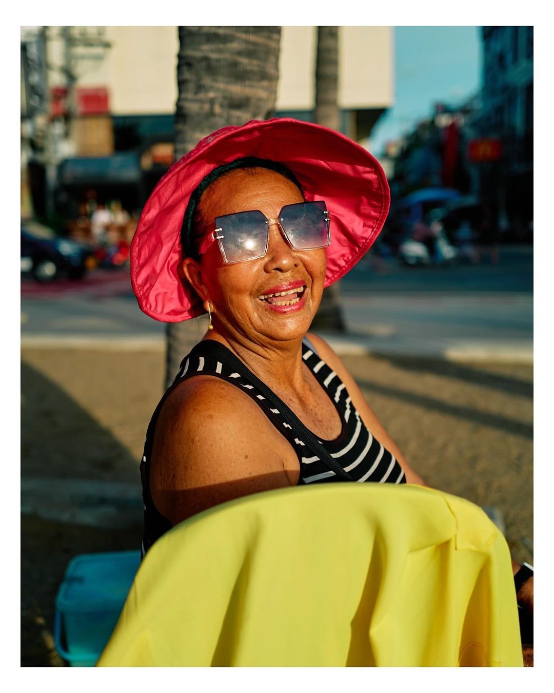 Smiling senior woman outdoors wearing a pink hat and sunglasses. She has a black and white striped top and a yellow garment.