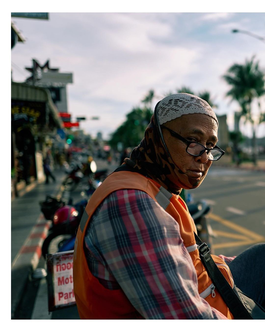 Portrait of an older adult woman with glasses and a headscarf, sitting outdoors in an urban setting with motorbikes and buildings.