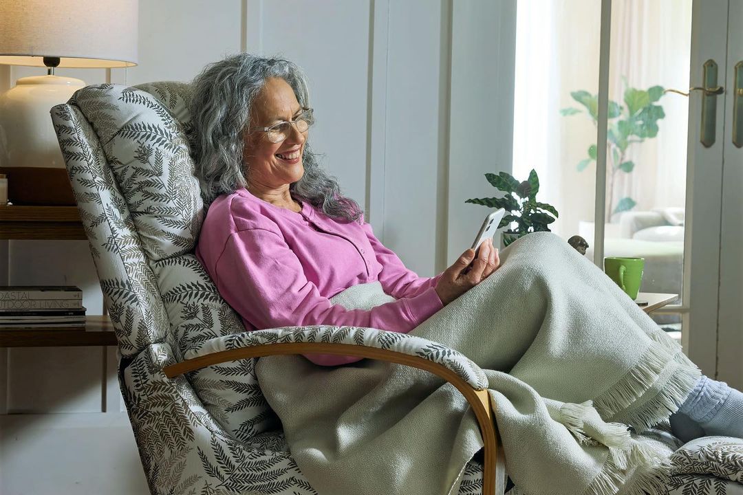 An older woman with grey hair and glasses relaxes in a patterned armchair while looking at her smartphone in a bright indoor space.