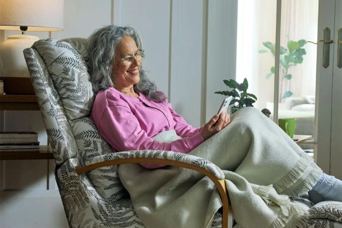 Elderly woman with grey hair smiling while looking at a smartphone, seated in a patterned armchair with a blanket.