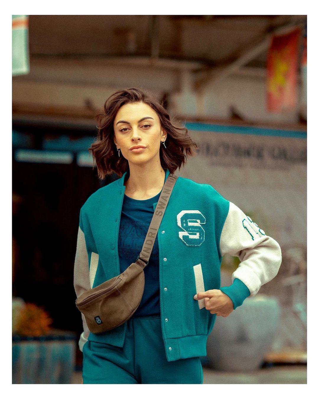Brunette woman in a teal letterman jacket and matching pants, wearing a brown fanny pack, poses outdoors with a neutral facial expression.