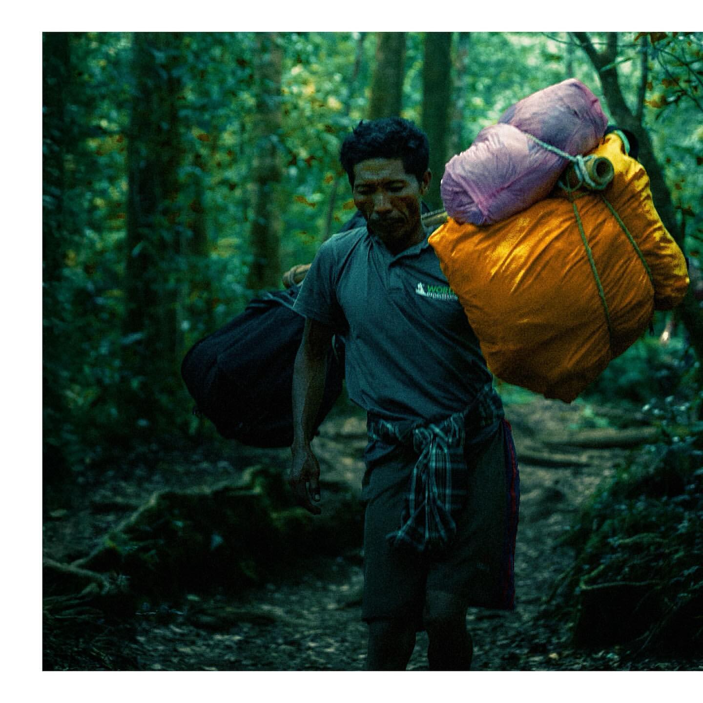 Man carrying heavy packs walks a path in a green forest. He wears casual clothes and a patterned wrap around his waist.