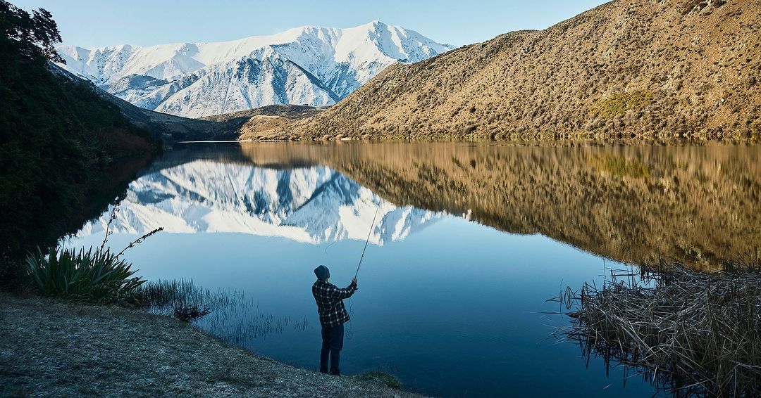 A man fly fishing in a tranquil lake with snow-capped mountains reflected in the water, wearing a flannel shirt and beanie.