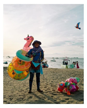Woman standing on a beach holding inflatable pool toys with a kite flying in the background on a sunny day near the ocean.