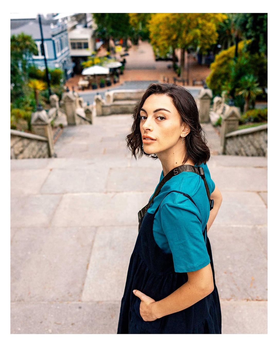 A young woman with short brown hair poses on stone stairs in an urban setting, wearing a blue top and dark dress. Buildings in the background.