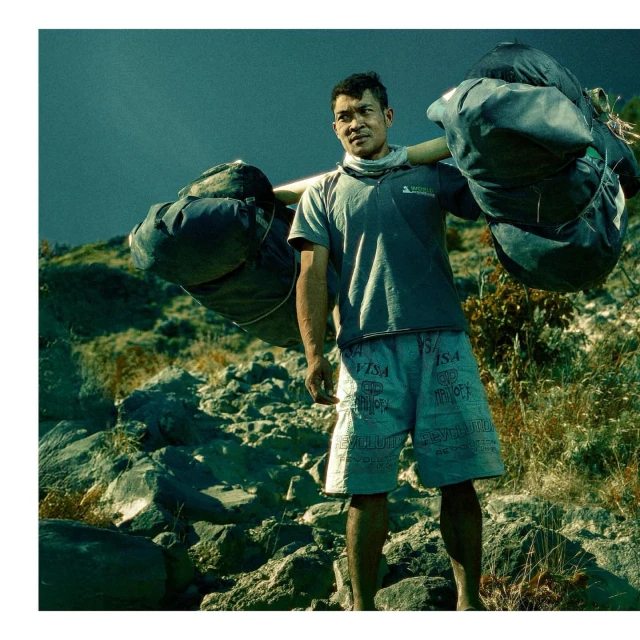 Asian man carries heavy sacks up rocky slope in outdoor shot with overcast lighting, creating a documentary-style image.