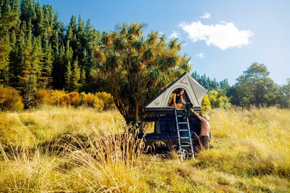 Two people with a tent atop a car in a grassy field with a backdrop of trees under a bright blue sky on a sunny day.
