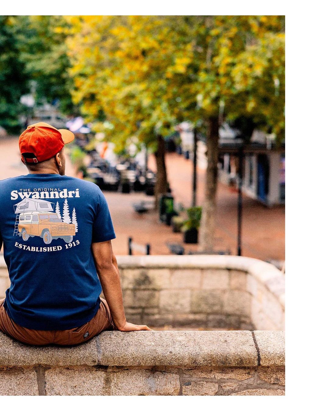 Man in navy t-shirt with truck graphic sitting on stone wall, looking at pedestrian street with trees and buildings on sunny day.