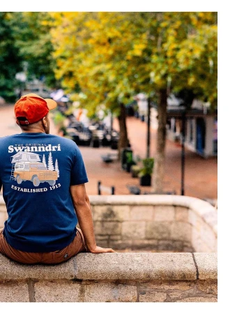 Man in navy t-shirt with truck graphic sitting on stone wall, looking at pedestrian street with trees and buildings on sunny day.