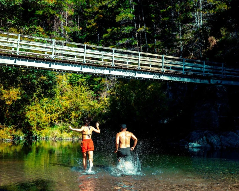 A man and woman are splashing in a shallow river beneath a pedestrian bridge surrounded by lush green trees in an outdoor, daytime scene.