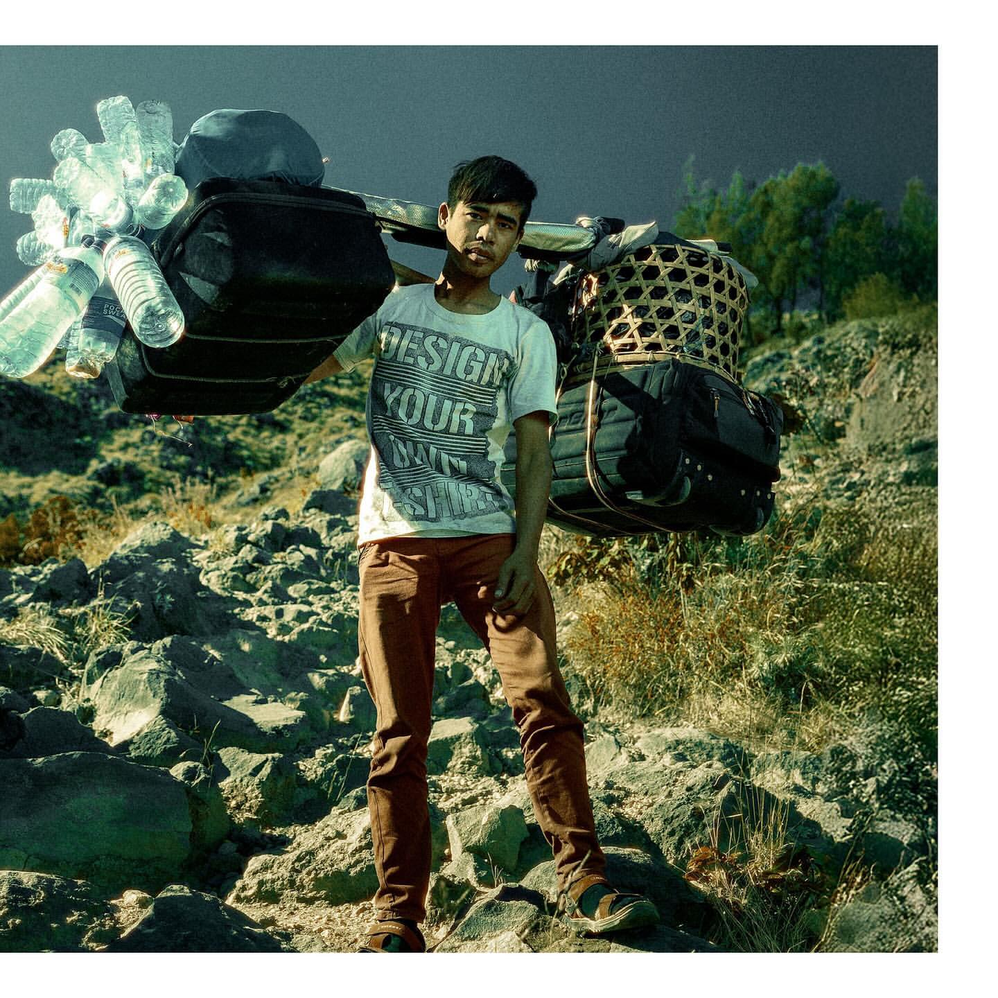 Full shot of a man carrying water bottles and a basket on his shoulders while standing on a rocky landscape with sparse vegetation.