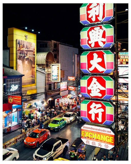 Night view of Bangkok, Thailand, featuring a busy street with cars and tuk-tuks, illuminated by vibrant neon signs and streetlights.