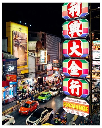 Night view of Bangkok, Thailand, featuring a busy street with cars and tuk-tuks, illuminated by vibrant neon signs and streetlights.