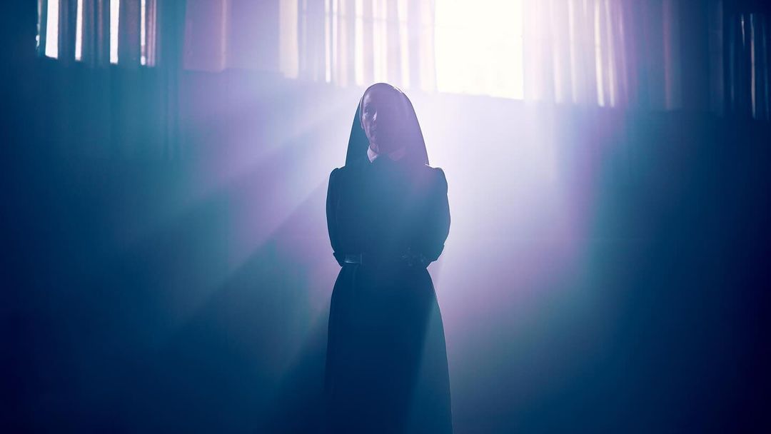 A single woman in a black religious habit stands in front of a bright window with rays of light beaming down in a dark room.