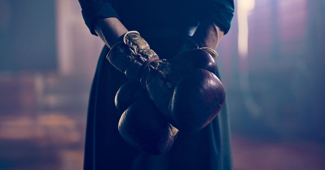 Close-up of a person holding worn boxing gloves tied together in a dimly lit setting, creating a moody, dramatic image.