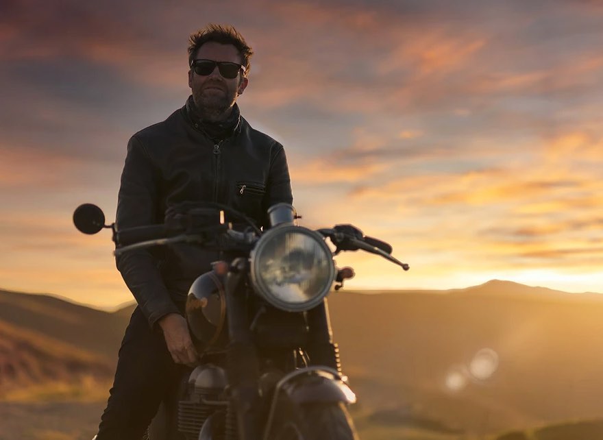 An adult man sits on a motorcycle in a black jacket and sunglasses against a warm sunset in a mountainous landscape.