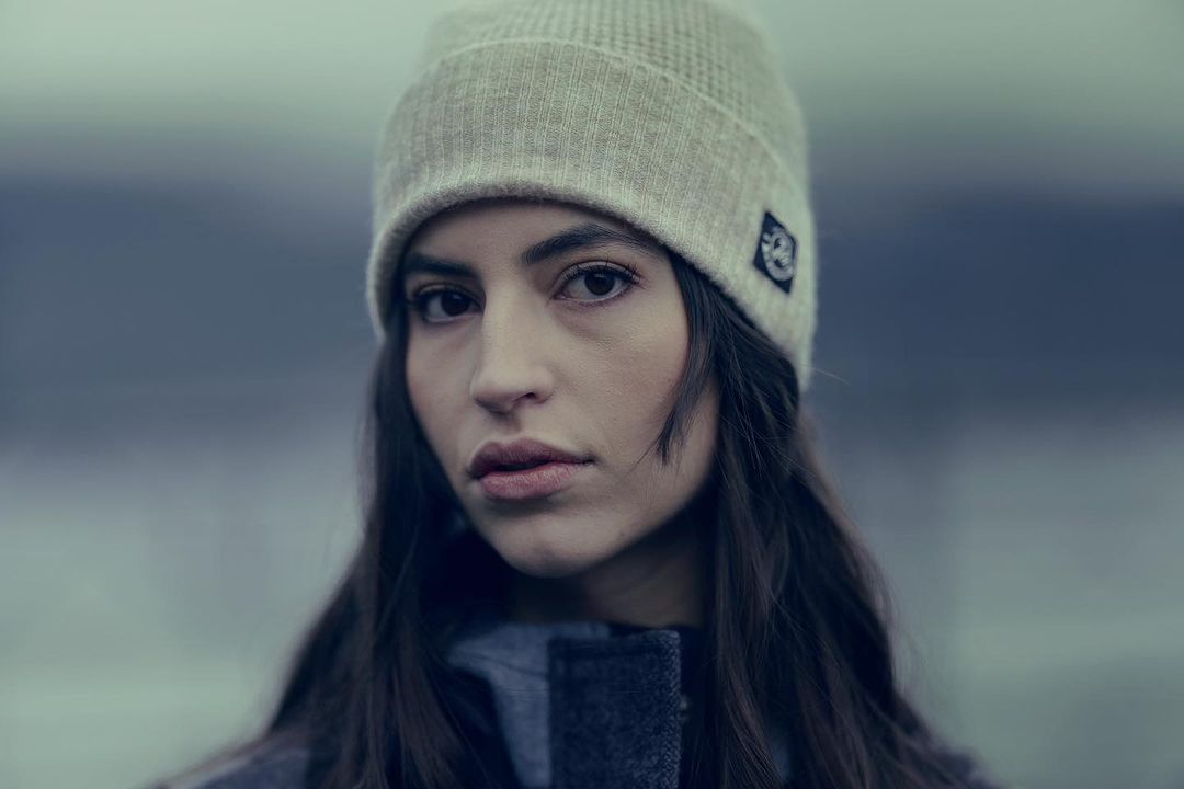 Portrait of a young woman wearing a knit beanie, looking directly at the camera in an outdoor setting with diffused lighting.