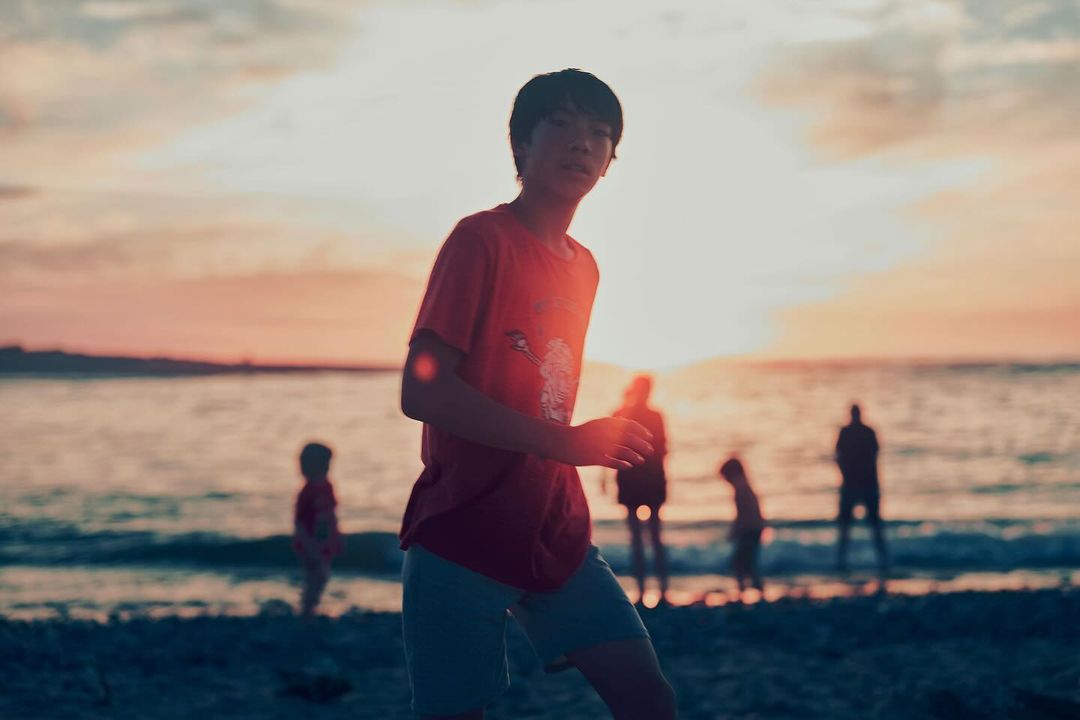 An adolescent boy stands on a beach at sunset with a group of silhouetted people near the water's edge.