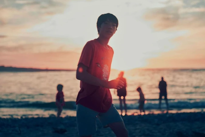 An adolescent boy stands on a beach at sunset with a group of silhouetted people near the water's edge.