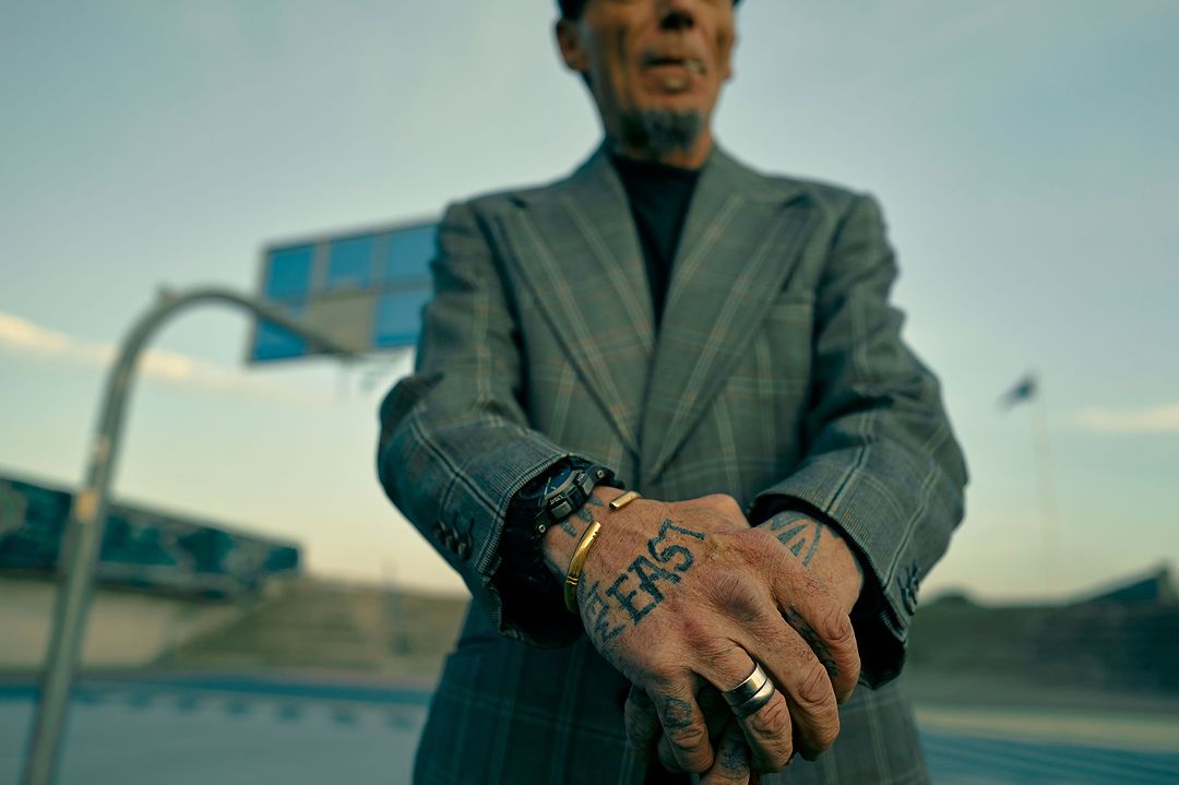Tattooed man in gray suit stands outdoors at basketball court, gold ring and bracelet, watch, hand tattoos, and gray patterned jacket.