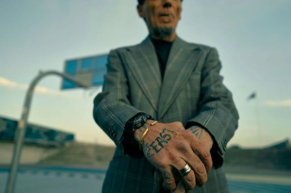Tattooed man in gray suit stands outdoors at basketball court, gold ring and bracelet, watch, hand tattoos, and gray patterned jacket.