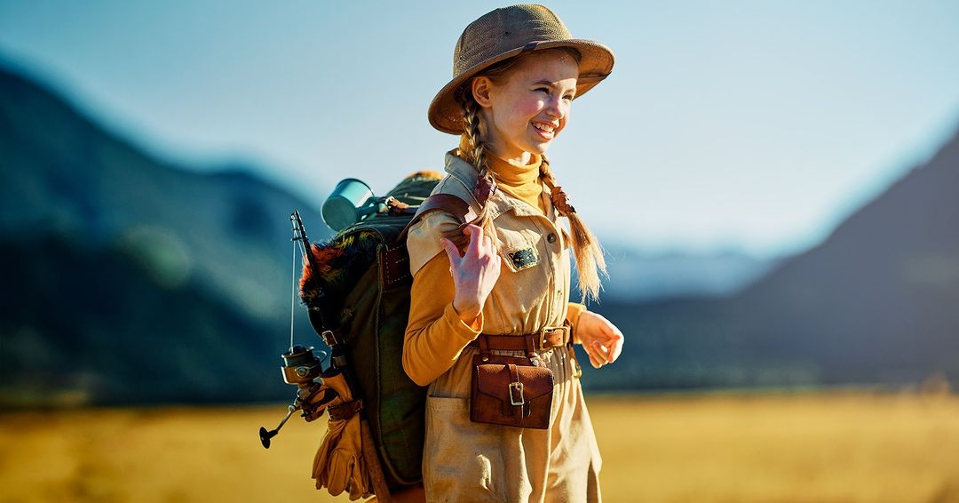 Young girl in safari outfit and hat, smiles while walking through a field with a backpack, fishing pole, and mountains in the background.