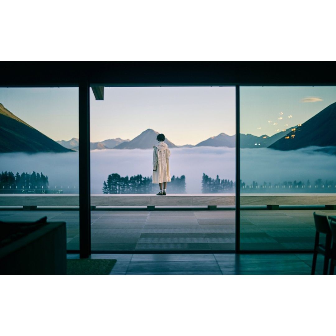 Person in a white coat standing on a balcony overlooking a mountain range covered in fog through a large window during daylight.