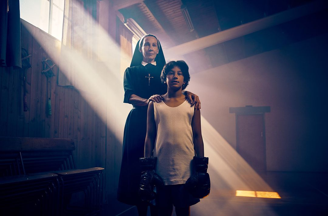 A nun in a black habit stands with a young boy wearing boxing gloves in a dimly lit room with light rays coming through the windows.