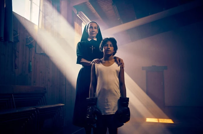 A nun in a black habit stands with a young boy wearing boxing gloves in a dimly lit room with light rays coming through the windows.