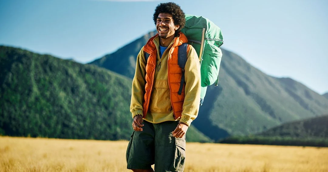 Smiling man hikes in outdoors wearing orange vest, backpack; set against mountain landscape with blue sky and golden grassy field.