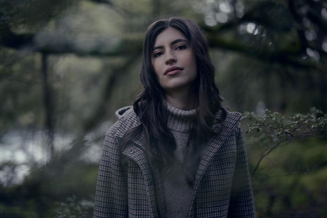 Portrait of a woman in a patterned coat standing outdoors in a forest, with a shallow depth of field and muted, natural colors.