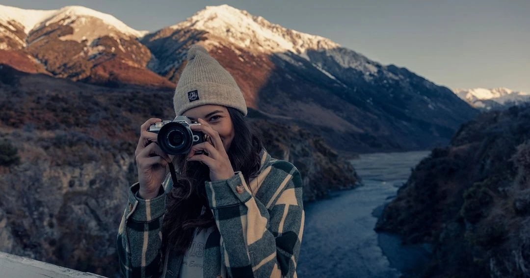 Young woman takes a picture with an analog camera against a background of snow-capped mountains and a river in the countryside.