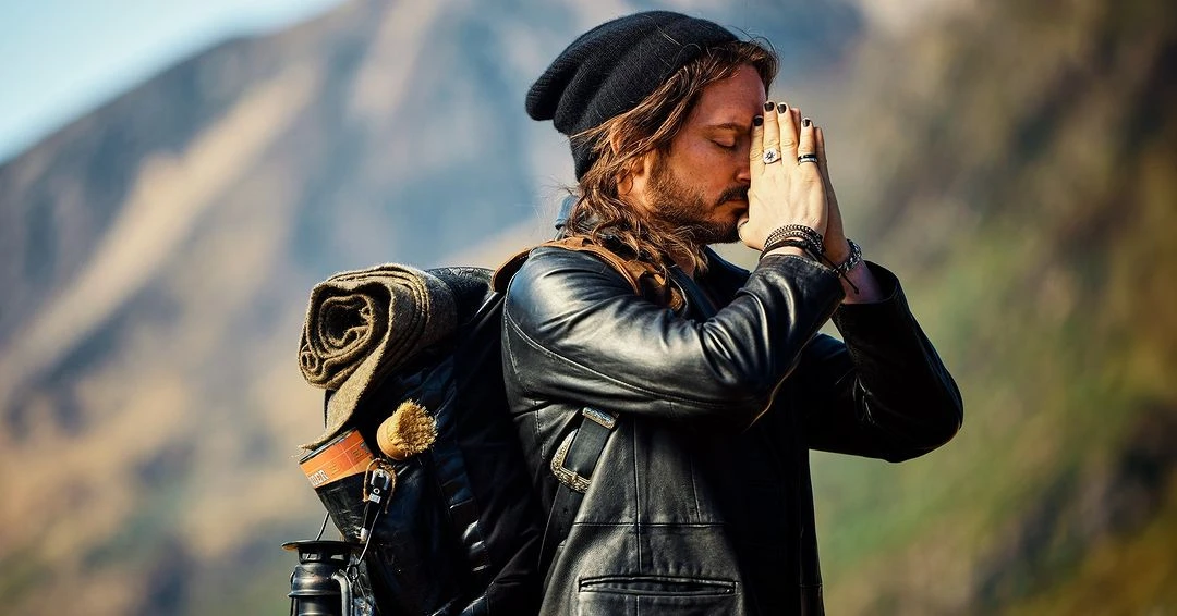 Adult male with a backpack standing in a mountainous landscape with his hands together, wearing a black beanie and a black jacket.