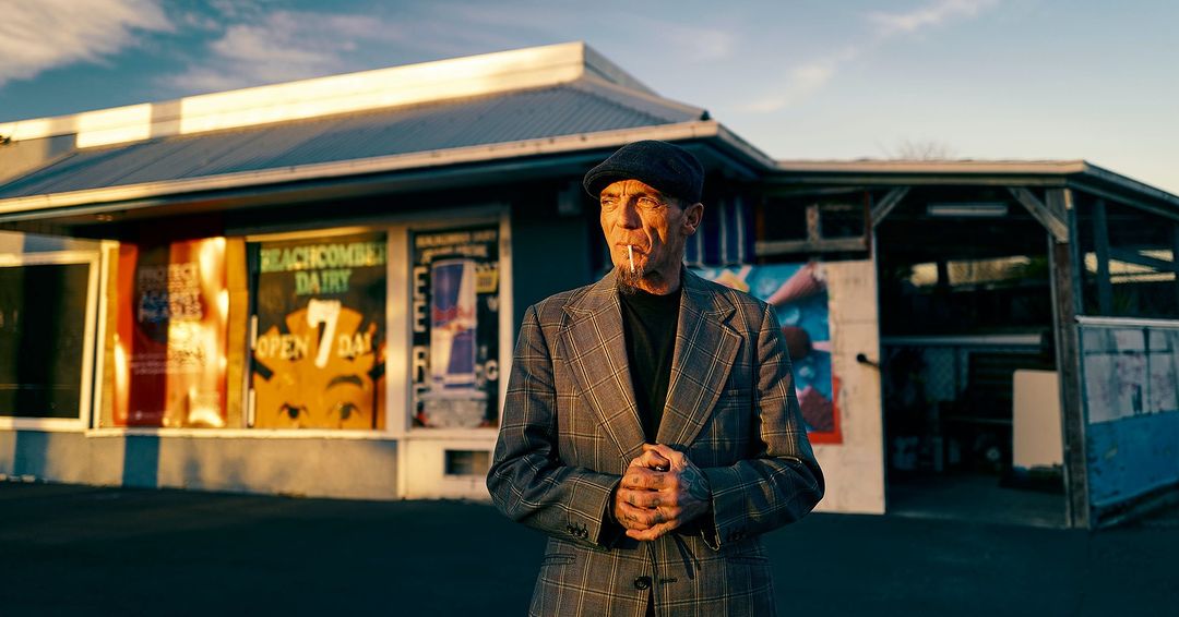Mature man in a grey plaid suit stands in front of a vintage dairy store with posters in the windows during golden hour sunlight.