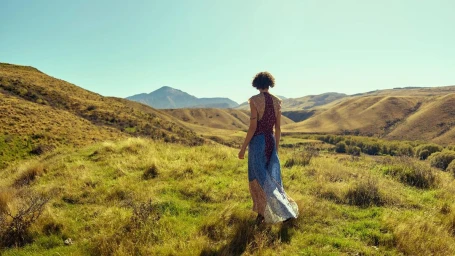 Model wearing a patterned maxi dress walking through a grassy hilly landscape under a clear sky.