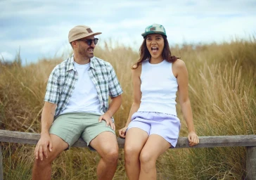 Man and woman sitting on a wooden fence in a grassy landscape wearing caps and casual summer clothing.
