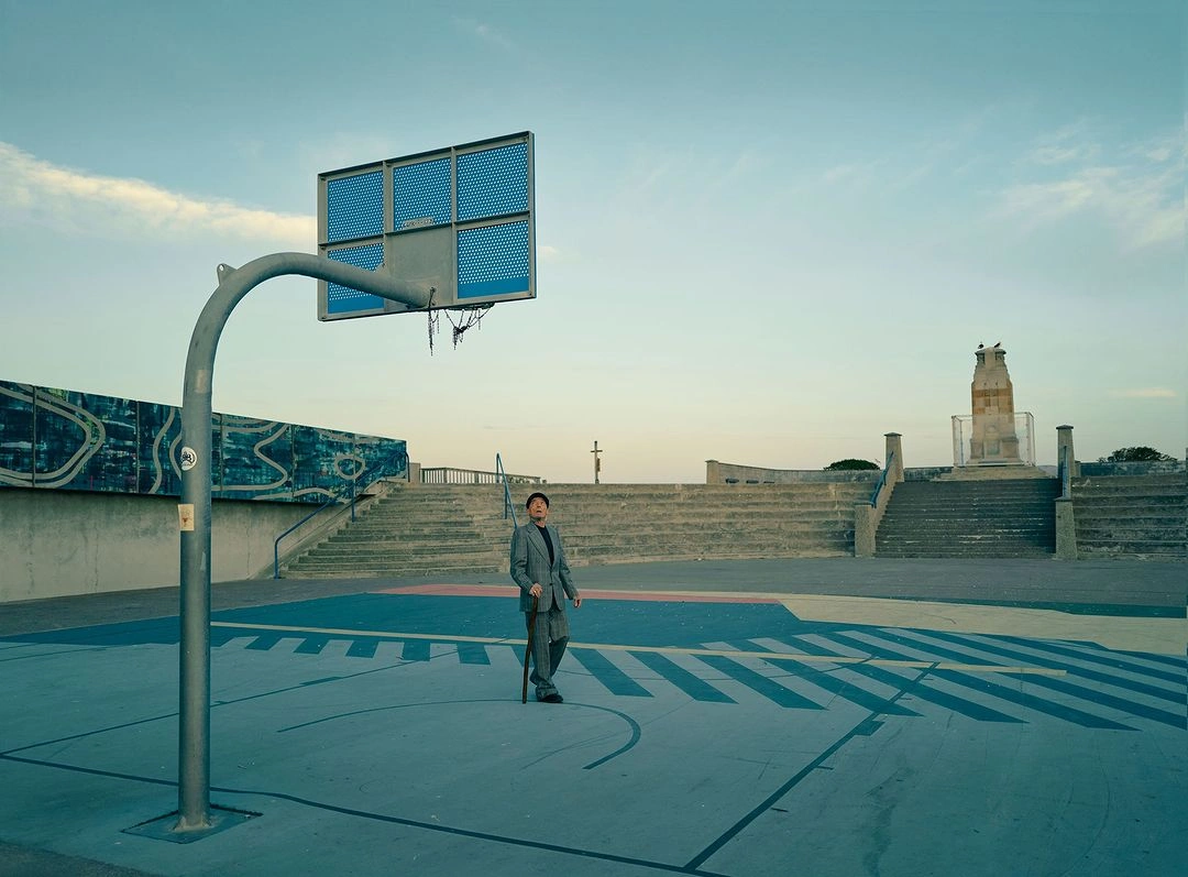 An elderly person in a grey suit holding a cane stands on a concrete basketball court beneath an outdoor hoop.