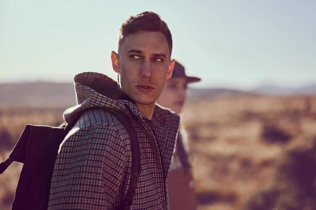 Man wearing a plaid hooded jacket and a backpack outdoors with another person blurred in the background
