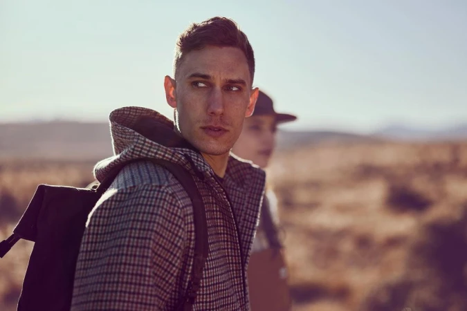 Man wearing a plaid hooded jacket and a backpack outdoors with another person blurred in the background