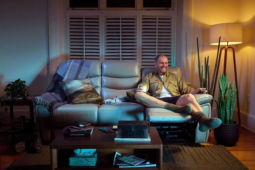 Man sitting on a grey leather couch holding a mug and remote control in a warmly lit living room by Dean Mackenzie