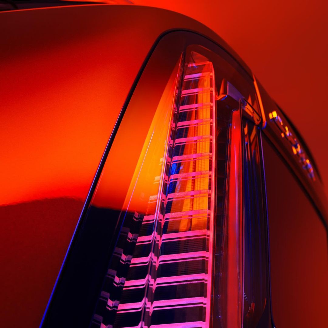 Close-up shot of an automotive tail light. The car body is sleek, reflecting vivid oranges and blues. The scene is high-contrast.