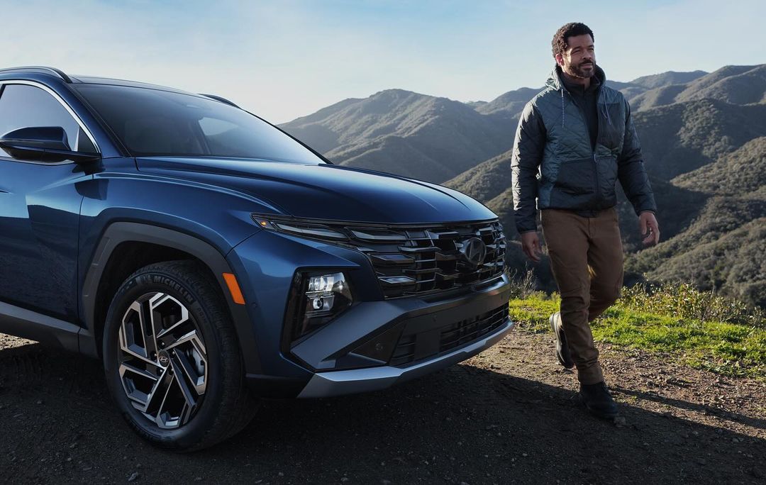 Dark blue SUV parked on a hillside with an adult male walking beside it, showcasing automotive design against a mountain backdrop.