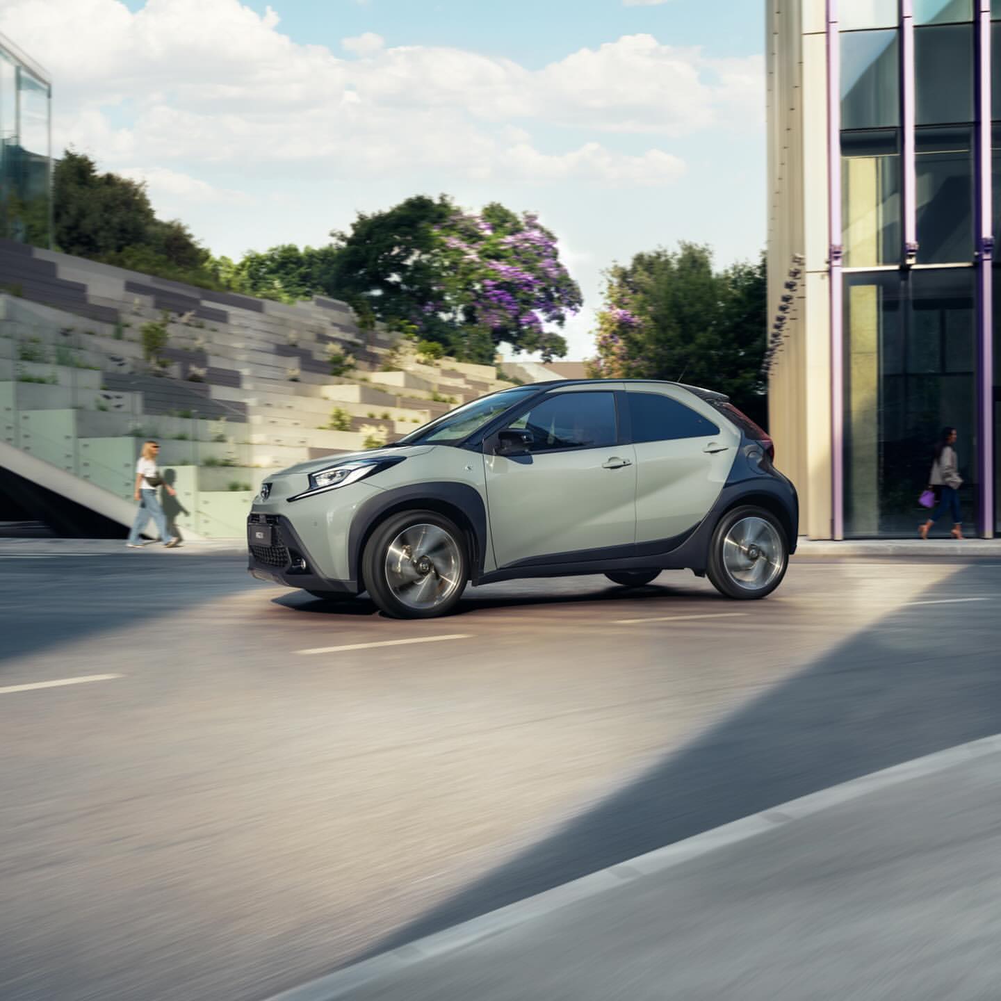 Silver compact car driving on a city street near a modern building and lush greenery under a bright sky on a sunny day.