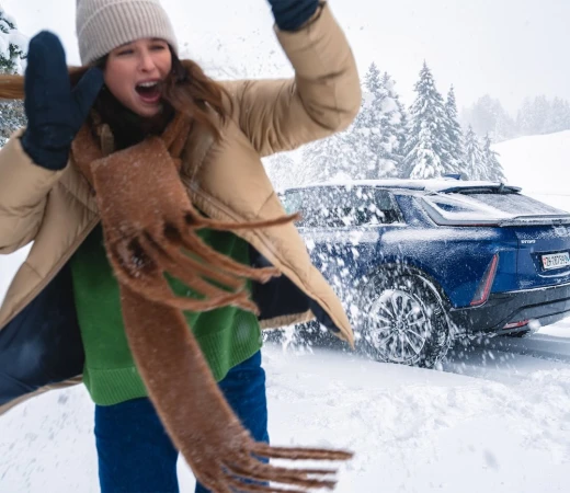 Excited woman in winter clothes throwing snow near a blue Cadillac Lyriq electric car in a snowy, wooded landscape on a cloudy day.