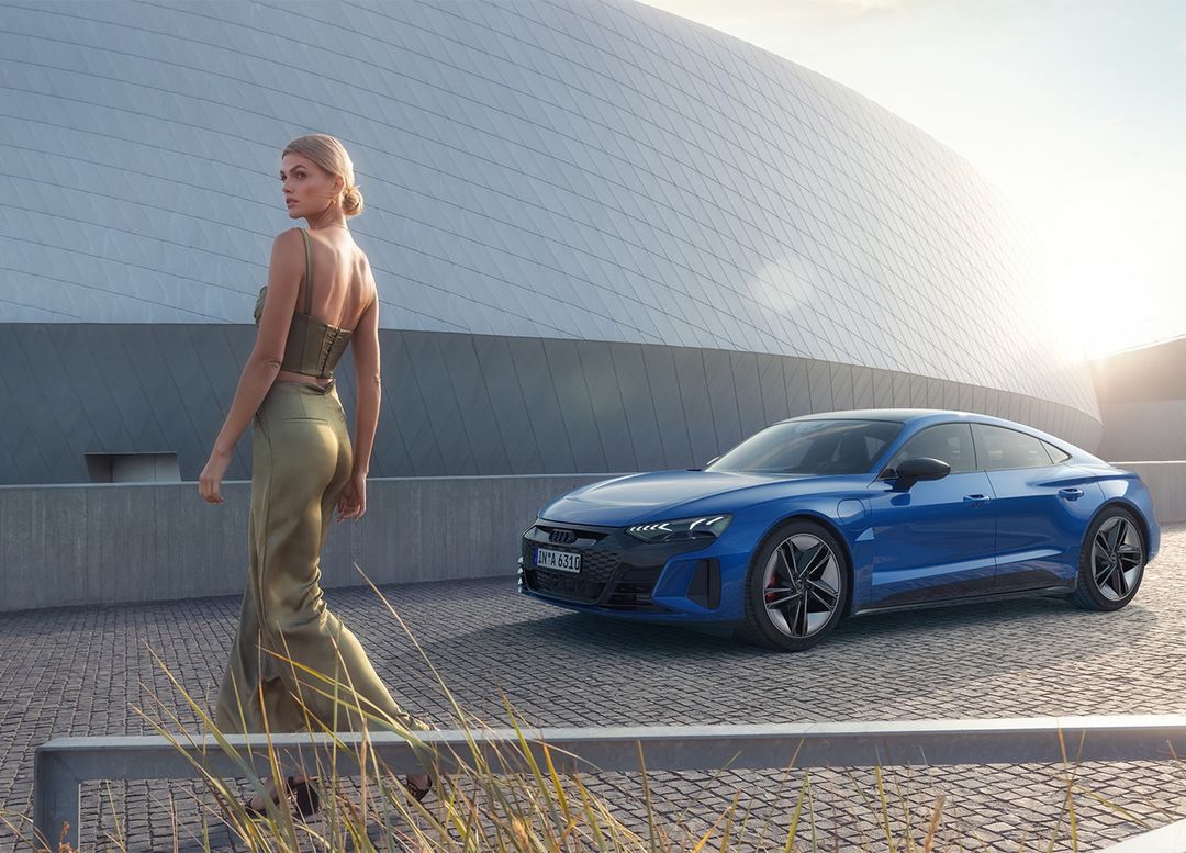 Adult female model in green satin outfit walking past a blue Audi e-tron GT parked in front of a modern architectural building.