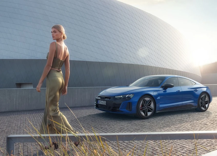 Adult female model in green satin outfit walking past a blue Audi e-tron GT parked in front of a modern architectural building.