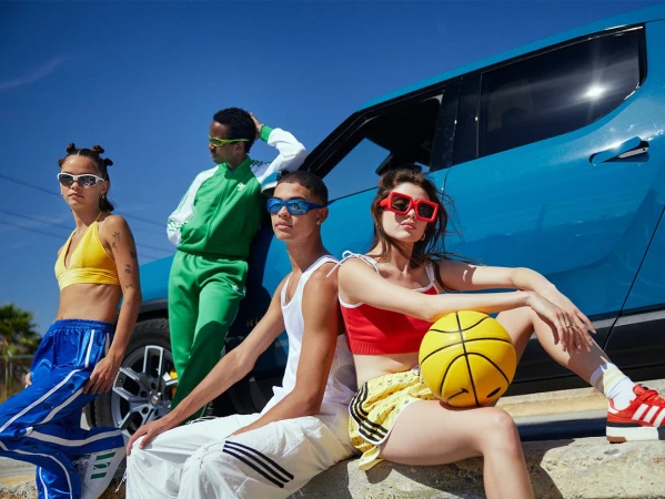 Group of four diverse young adult models wearing sports-inspired clothing and sunglasses, posed in front of a bright blue car on a sunny day.